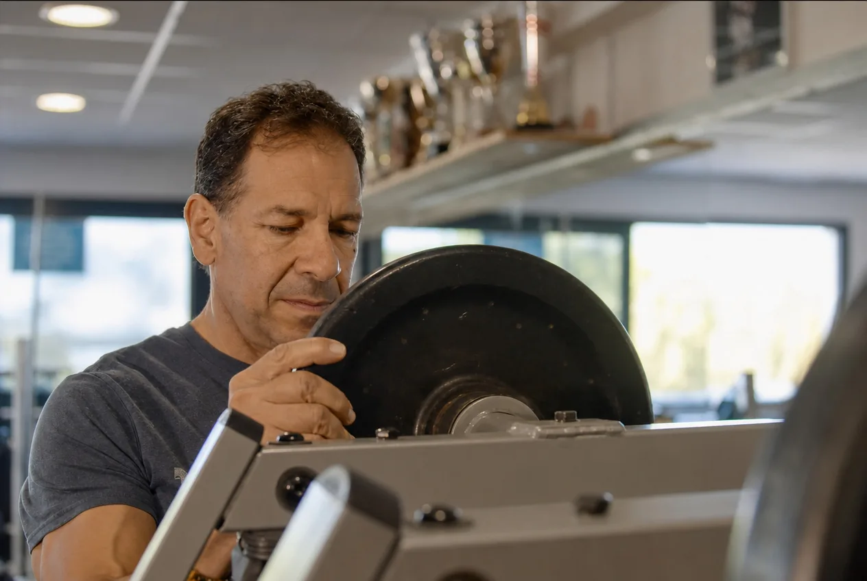 Adhérent d'ABS Sport Gym chargeant un disque sur une machine, trophées du club en arrière-plan